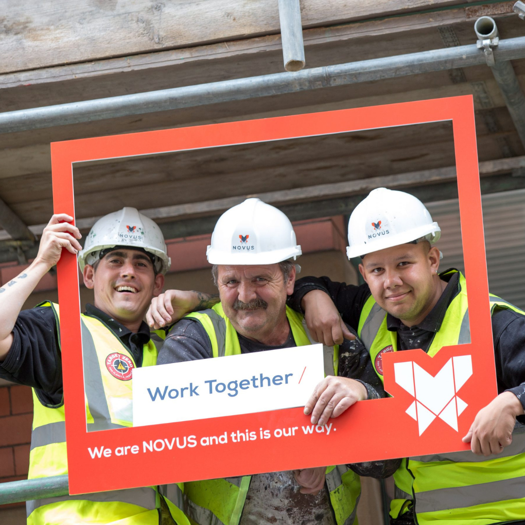 Three men on a scaffolding platform holding a frame which states "Work together, we are NOVUS and this is our way"