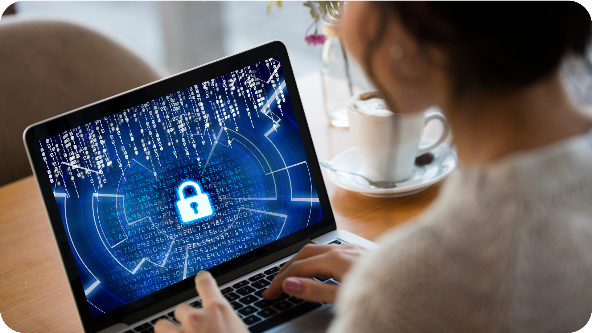 Close-up of a laptop screen displaying a white lock icon on a blue digital background with binary numbers, symbolizing cybersecurity and data protection, with hands typing on the keyboard and a coffee cup on the table.